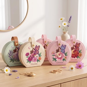 Peut inclure: Cinq boîtes à bijoux rondes de couleur pastel avec poignées supérieures et fermetures à pression. Chaque boîte présente une illustration imprimée d'une fée avec des ailes, des fleurs, un croissant de lune et le nom Vivienne en écriture cursive. Les boîtes sont vert clair, crème et dans des tons roses.