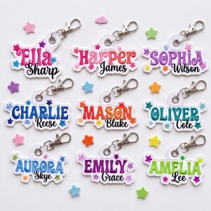 Puede incluir: Nueve llaveros bordados personalizados con cierres de langosta plateados, cada uno con un nombre en letras coloridas de estilo inflado rodeado de pequeñas estrellas bordadas. Los nombres son Ella Sharp, Harper James, Sophia Wilson, Charlie Reese, Mason Blake, Oliver Cole, Aurora Skye, Emily Grace y Amelia Lee.