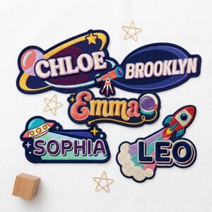 May include: Five embroidered patches featuring space-themed designs and names. The patches include a planet with 'CHLOE', a dark oval with 'BROOKLYN', a colorful script 'Emma', a UFO with 'SOPHIA', and a rocket ship with 'LEO'. Gold wire stars are scattered.