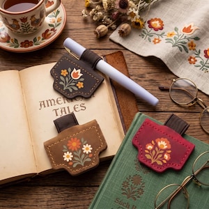 May include: Three leather pen holders with elastic bands, each featuring a unique floral print. One is dark brown with a tulip, one is light brown with daisies, and one is red with a flower cluster. They are placed on open books.