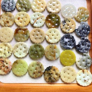 May include: A selection of circular stone ornaments, each with a cracker-like design and multiple colours. The pieces are displayed on a wooden tray, with shades of beige, green, and brown. The ornaments have a variety of textures.