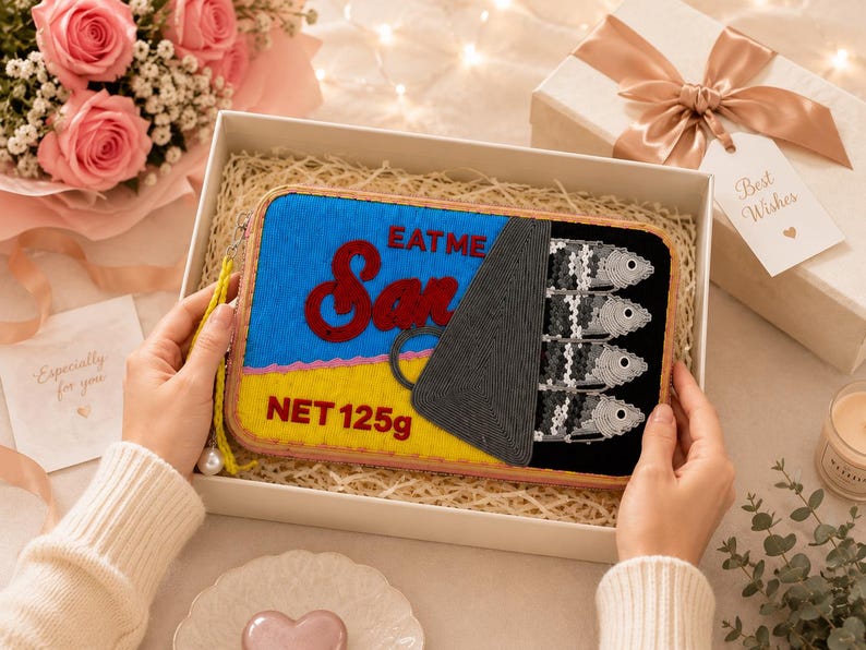 May include: A rectangular zippered pouch designed to resemble a sardine tin. The front features blue and yellow sections with red text reading EAT ME and NET 125g, alongside a black section depicting four embroidered silver fish and a pull tab.