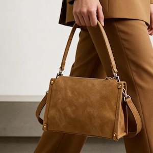 May include: A tan suede handbag with a rectangular shape. The bag features a top handle and a detachable shoulder strap. The bag has silver-coloured hardware. The bag is being held by a person wearing a brown suit.
