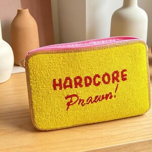 May include: A rectangular yellow beaded clutch bag featuring the red embroidered text HARDCORE Prawn! on the front. The bag has a pink zipper closure along the top edge and is finished with a metallic gold trim around the perimeter of the pouch.