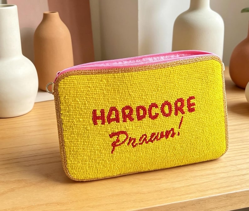 May include: A rectangular yellow beaded clutch bag featuring the red embroidered text HARDCORE Prawn! on the front. The bag has a pink zipper closure along the top edge and is finished with a metallic gold trim around the perimeter of the pouch.