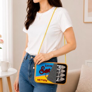 May include: A rectangular crossbody bag shaped like a sardine tin, featuring a yellow base, blue top, and a grey peel-back lid. The bag displays the text "EAT ME" and "NET 125g" with a graphic of four silver sequined fish on the right side.