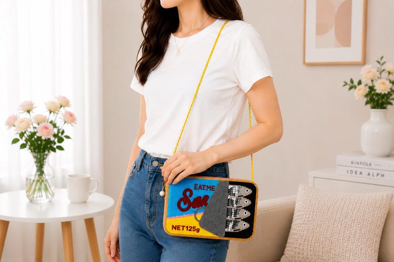 May include: A rectangular crossbody bag shaped like a sardine tin, featuring a yellow base, blue top, and a grey peel-back lid. The bag displays the text "EAT ME" and "NET 125g" with a graphic of four silver sequined fish on the right side.
