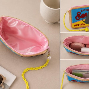 May include: A rectangular zippered pouch featuring a sardine tin graphic with the text EAT ME Sardines NET 125g. The exterior has a blue, yellow, and black design, while the interior is lined with pink satin. It includes a yellow braided wrist strap.