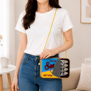 May include: A rectangular crossbody bag designed to look like a sardine tin. The bag features a blue and yellow label with the text "EAT ME" and "Sardines" in red script, a grey peel-back lid, and a graphic of four silver fish. It has a thin yellow strap.