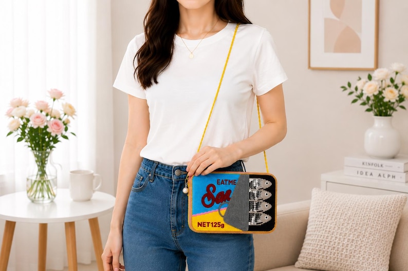 May include: A rectangular crossbody bag designed to look like a sardine tin. The bag features a blue and yellow label with the text "EAT ME" and "Sardines" in red script, a grey peel-back lid, and a graphic of four silver fish. It has a thin yellow strap.