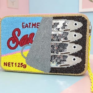 May include: A rectangular beaded crossbody bag designed to look like a sardine tin. The front features light blue, yellow, and black beads with red text reading EAT ME and Sardines, plus NET 125g. Four silver sequined fish are visible.