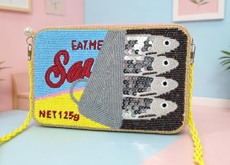 May include: A rectangular beaded crossbody bag designed to look like a sardine tin. The front features light blue, yellow, and black beads with red text reading EAT ME and Sardines, plus NET 125g. Four silver sequined fish are visible.