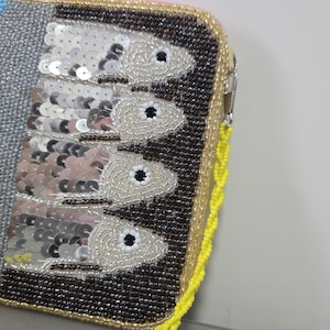 May include: A rectangular zippered clutch featuring intricate beadwork and sequins. The design displays four fish arranged vertically against a dark brown beaded background, with a silver metallic section and a bright yellow beaded border trim.