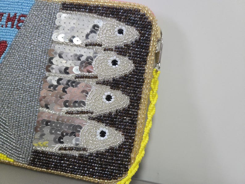 May include: A rectangular zippered clutch featuring intricate beadwork and sequins. The design displays four fish arranged vertically against a dark brown beaded background, with a silver metallic section and a bright yellow beaded border trim.