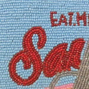 May include: A rectangular beaded pouch featuring a light blue background with the red text EAT ME and the word Sad in a larger red script font. The edges are trimmed with gold beads, and a section of silver beads is visible on the right side.
