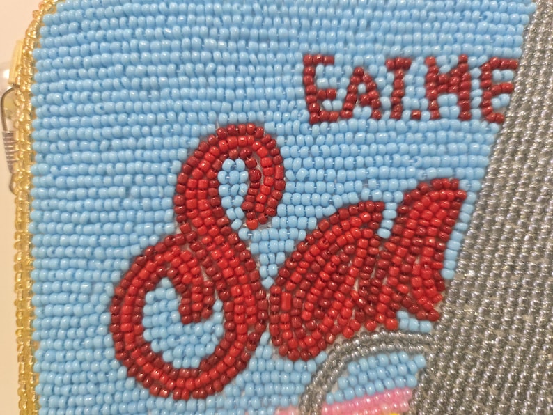 May include: A rectangular beaded pouch featuring a light blue background with the red text EAT ME and the word Sad in a larger red script font. The edges are trimmed with gold beads, and a section of silver beads is visible on the right side.