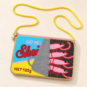 May include: A rectangular beaded crossbody bag shaped like a shrimp can. The design features a blue and yellow background with red text reading EAT ME and Shrimp, and black text NET 125g. It includes a silver lid graphic and pink shrimp.