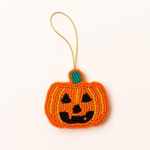 May include: A pumpkin-shaped ornament crafted from small orange, yellow, and black beads. The jack-o'-lantern features a teal bead stem and a gold-coloured hanging loop. The facial features, including triangular eyes and a jagged mouth, are formed by black beads.