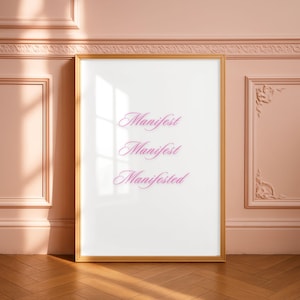 May include: A framed art print with the words "Manifest, Manifest, Manifested" in a cursive font. The print has a gold frame and is leaning against a pink wall with decorative molding. The floor is made of wood.