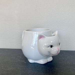 Może przedstawiać: Biały ceramiczny kubek w kształcie kota. Kubek ma szczeg&oacute;łową kocią twarz z r&oacute;żowymi akcentami na uszach i nosie, czarnymi oczami i wąsami. Kubek ma uchwyt i jest przeznaczony do stania.