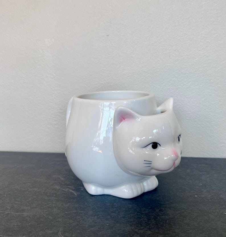 Może przedstawiać: Biały ceramiczny kubek w kształcie kota. Kubek ma szczeg&oacute;łową kocią twarz z r&oacute;żowymi akcentami na uszach i nosie, czarnymi oczami i wąsami. Kubek ma uchwyt i jest przeznaczony do stania.