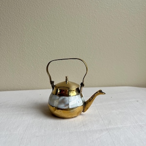 May include: A small brass teapot featuring a central band of iridescent mother-of-pearl tiles. The teapot has a polished brass lid, a curved brass spout, and a hinged brass handle that arches over the top of the rounded metal body.