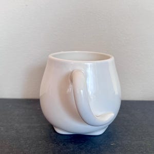 Może przedstawiać: Biały ceramiczny kubek o zaokrąglonym korpusie i zakrzywionym uchwycie. Kubek ma błyszczące wykończenie i stoi na ciemnej powierzchni. Kubek jest przeznaczony do przechowywania gorących lub zimnych napoj&oacute;w.