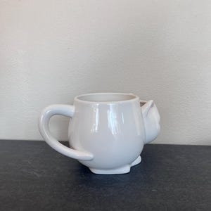 Może przedstawiać: Biały ceramiczny kubek w kształcie kota, z uchwytem i detalem w postaci kociej twarzy z boku. Kubek ma błyszczące wykończenie i stoi na ciemnej powierzchni. Przeznaczony do picia napoj&oacute;w.