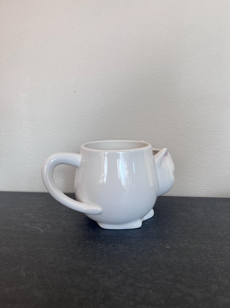 Może przedstawiać: Biały ceramiczny kubek w kształcie kota, z uchwytem i detalem w postaci kociej twarzy z boku. Kubek ma błyszczące wykończenie i stoi na ciemnej powierzchni. Przeznaczony do picia napoj&oacute;w.