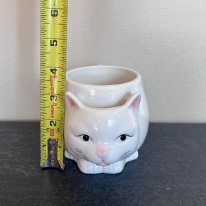 Może przedstawiać: Biały ceramiczny kubek z czarnym uchwytem i napisem "But First, Coffee" czarną, pogrubioną czcionką. Kubek stoi na białej powierzchni. Kubek ma 10 cm wysokości.