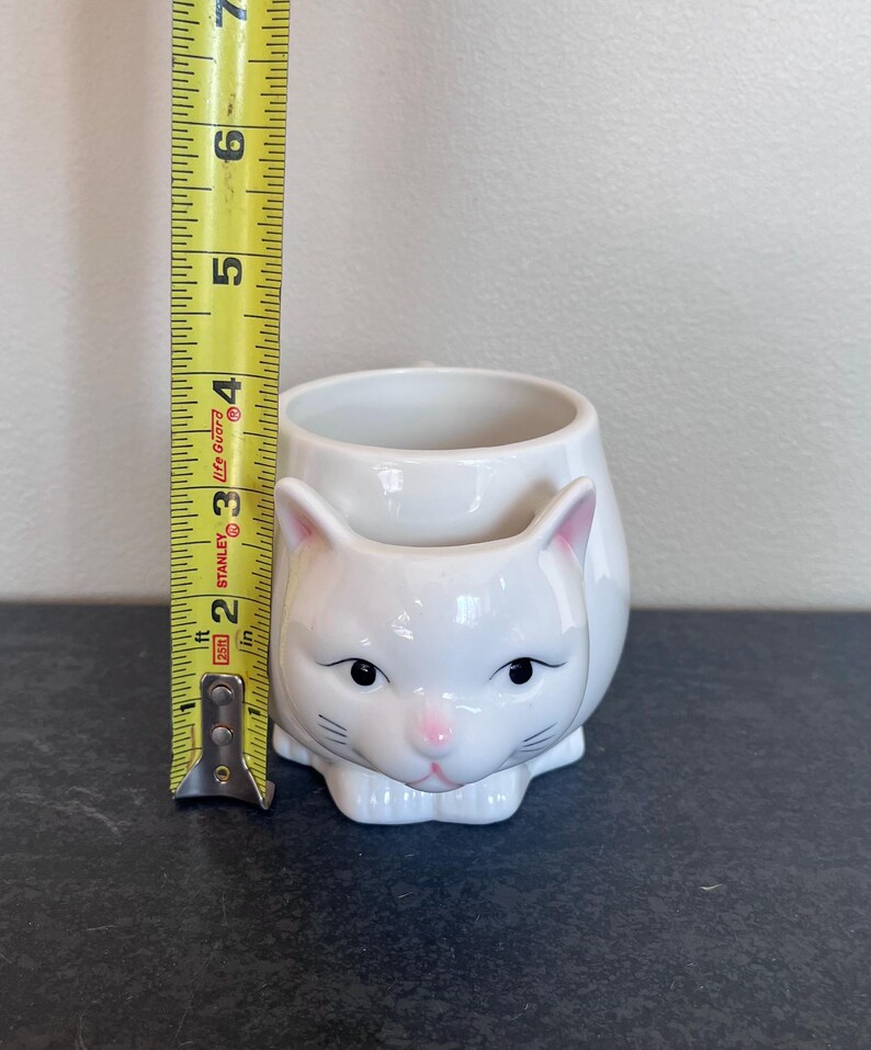 Może przedstawiać: Biały ceramiczny kubek z czarnym uchwytem i napisem "But First, Coffee" czarną, pogrubioną czcionką. Kubek stoi na białej powierzchni. Kubek ma 10 cm wysokości.