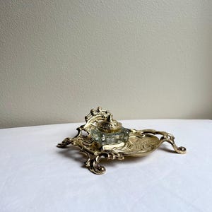 May include: A gold-coloured metal inkwell stand featuring an ornate, swirling design with four curved feet. A clear, square glass ink container with a matching gold-coloured metal lid sits securely in the centre of the decorative metal base.