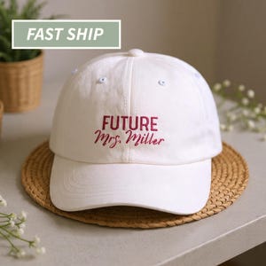 May include: A white baseball cap featuring the embroidered text "FUTURE Mrs. Miller" in burgundy. The cap has a curved peak and a classic six-panel design. The cap is on a woven coaster.