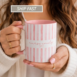 May include: A ceramic mug with a pink and white vertical striped pattern. The mug features the text nurse emely in a cursive script font with a small bow icon underneath. The interior of the mug and the handle are solid light pink.