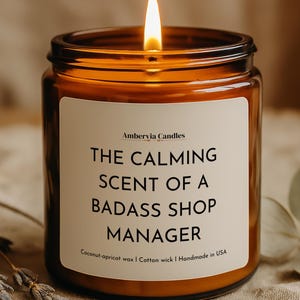 Puede incluir: Una vela encendida en un frasco de vidrio ámbar con una etiqueta blanca que presenta el texto Ambervia Candles, THE CALMING SCENT OF A BADASS SHOP MANAGER, Coconut-apricot wax, Cotton wick y Handmade in USA, con una llama en la parte superior.