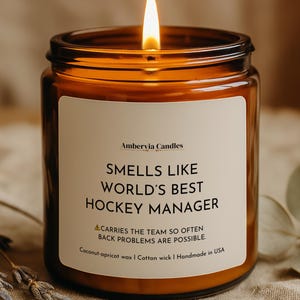 May include: A lit candle in an amber glass jar with a white label that reads: Ambervia Candles, SMELLS LIKE WORLD'S BEST HOCKEY MANAGER, CARRIES THE TEAM SO OFTEN BACK PROBLEMS ARE POSSIBLE, Coconut-apricot wax | Cotton wick | Handmade in USA.