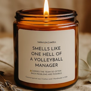 May include: A lit candle in an amber glass jar with a white label that reads: Ambervia Candles, SMELLS LIKE ONE HELL OF A VOLLEYBALL MANAGER, CARRIES THE TEAM SO OFTEN BACK PROBLEMS ARE POSSIBLE, Coconut-apricot wax | Cotton wick | Handmade in USA.
