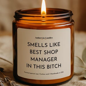 May include: A lit candle in an amber glass jar with a white label that reads Ambervia Candles, SMELLS LIKE BEST SHOP MANAGER IN THIS BITCH, Coconut-apricot wax | Cotton wick | Handmade in USA. The candle features a single burning wick.