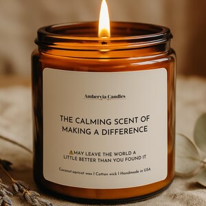 May include: A lit candle in an amber glass jar with a white label. The label reads Ambervia Candles, The Calming Scent of Making a Difference, May Leave the World a Little Better Than You Found It, Coconut-apricot wax, Cotton wick, Handmade in USA.