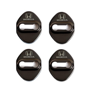 May include: Four black car door lock covers with the Honda logo in silver. Each cover is a dark, glossy color with a rounded, diamond-like shape and a keyhole opening. The Honda logo and text are centered.