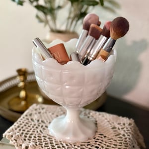 May include: A white milk glass pedestal bowl with a scalloped rim, holding makeup brushes and a tube of lipstick. The bowl has a diamond pattern and sits on a crocheted doily. A decorative item for a vanity or bathroom.