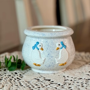 May include: A white ceramic planter with a speckled blue pattern. Two embossed white geese with blue bows and yellow feet decorate the sides. The planter has a rounded shape and a wide opening.