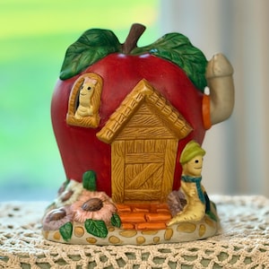 May include: A whimsical ceramic apple-shaped house with a red body, green leaves, and a brown stem. The house features a wooden door, a chimney, and a small window with a smiling character. A small worm figure is seated outside the door.