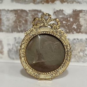 May include: A round gold-toned metal picture frame featuring an ornate embossed border with leaf patterns and a decorative bow at the top. The frame has a circular center opening and a textured metallic finish throughout the entire structure.