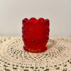May include: A small, red glass votive candle holder with a scalloped rim and a textured, starburst pattern. The base has a yellow tint. The candle holder is approximately 7.6 cm tall.