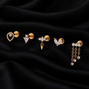 Könnte beinhalten: Fünf goldfarbene Ohrstecker mit flacher Rückseite und klaren Zirkonia-Steinen. Die Designs umfassen einen perlenbesetzten Tropfen, ein Dreieck, eine Marquise-Form mit Anhänger, ein blattartiges Element und drei Kettenanhänger.