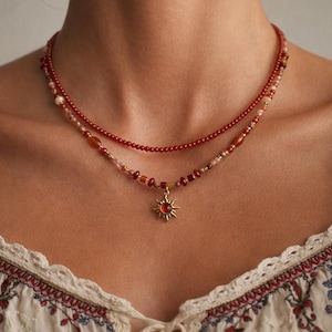 May include: A layered necklace featuring two strands of beads in shades of red, orange, and pink. The lower strand has a gold sun pendant with a red center. The necklace is worn against a light background.