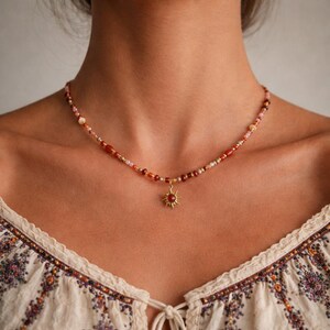 May include: A beaded necklace with a sun pendant. The necklace features a mix of red, orange, and gold beads. The sun pendant is gold-colored with a red center. The necklace is worn around the neck.