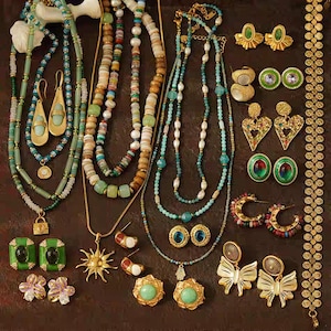 Puede incluir: Una colección de joyas vintage, que incluye collares, pendientes y pulseras. Los collares presentan cuentas de varios colores, como turquesa, blanco y dorado. Los pendientes incluyen aros dorados, tachuelas y estilos colgantes. Las joyas están dispuestas sobre una superficie marrón oscuro.
