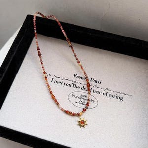 May include: A beaded necklace with a sun pendant. The necklace features small, colorful beads in shades of red, orange, and pink. The sun pendant is gold-colored with a red center. The necklace is displayed on a white surface with text.